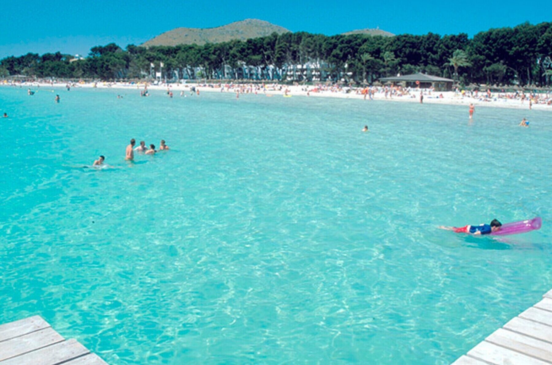 Ein Strand mit klarem türkisblauem Wasser, Menschen beim Schwimmen und Sonnenbaden sowie ein Holzsteg.