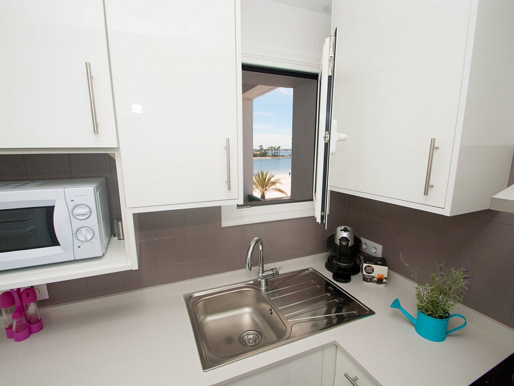 A modern kitchen with white cabinets, a stainless steel sink, a microwave, a coffee maker, and a window with a view of a beach.