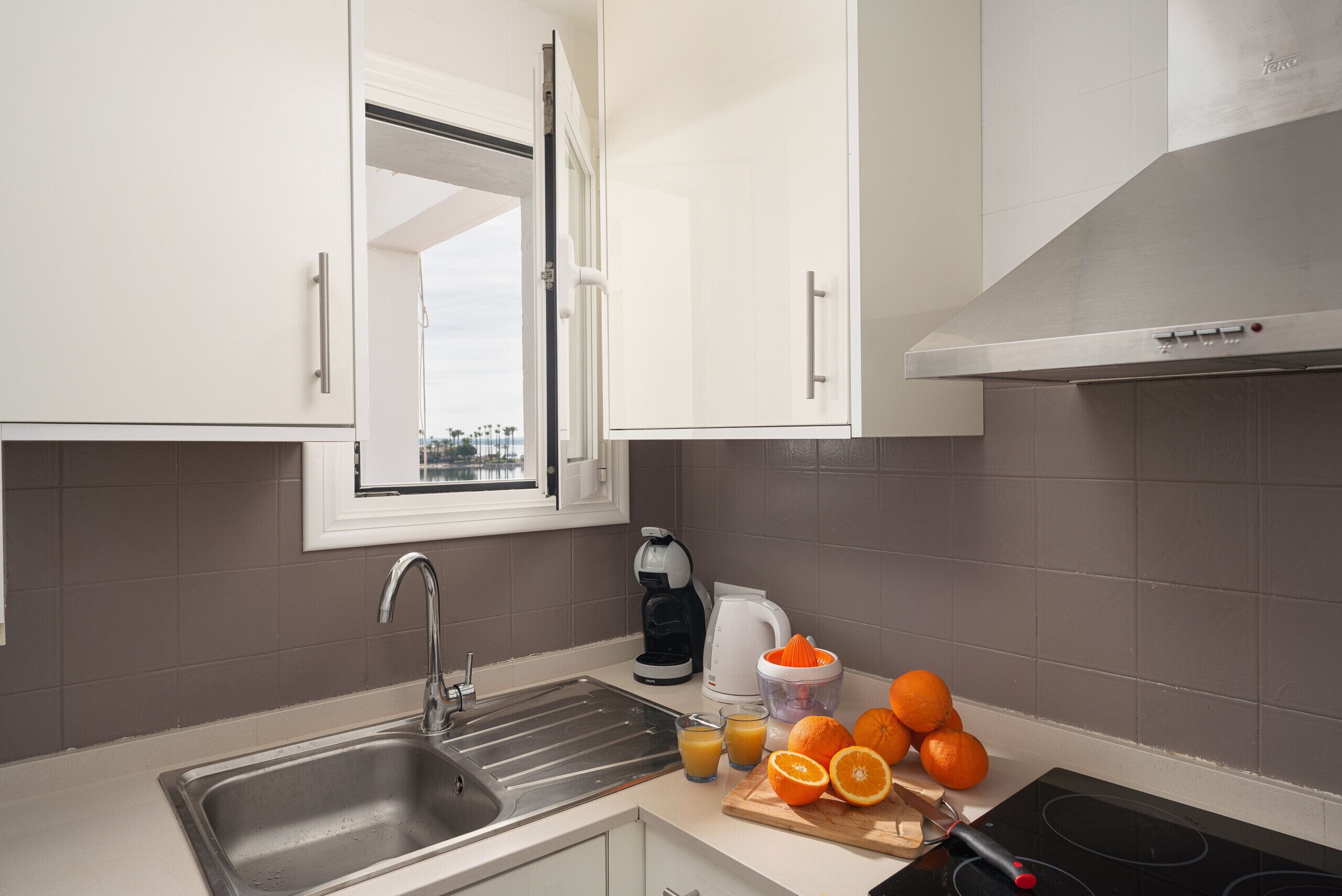 A modern kitchen with a stainless steel sink, a coffee maker, a kettle, and a cutting board with oranges.