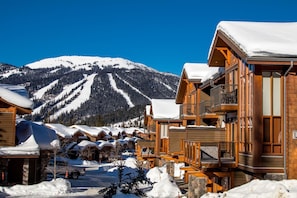 A snow-covered lodge with wooden architecture, a ski slope in the background, and a clear blue sky.
