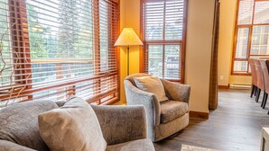 A cozy living room with a grey armchair, a wooden floor, and large windows with blinds. - Spacious multi level 4 bedroom + den townhouse, close to ski run, great views of Mt. Morrisey (Sun Peaks)