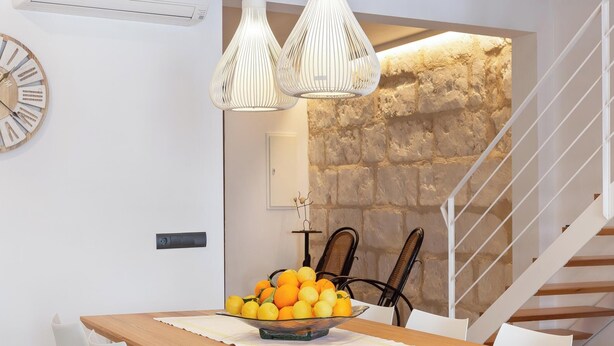 Ein moderner Essbereich mit einem Holztisch, weißen Stühlen und einer Schüssel Orangen. Im Hintergrund ist eine Treppe mit weißer Geländer und eine Steinsäule zu sehen.