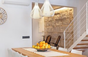 Un espace repas moderne avec une table en bois, des chaises blanches et un saladier rempli d’oranges. On aperçoit un escalier avec une rampe blanche et, au fond, un mur en pierre.