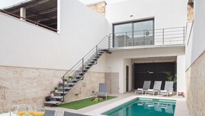 Ein moderner Poolbereich im Freien mit Glasgeländer, einer Treppe zu einer erhöhten Ebene und einer Lounge-Bereich mit Liegestühlen.
