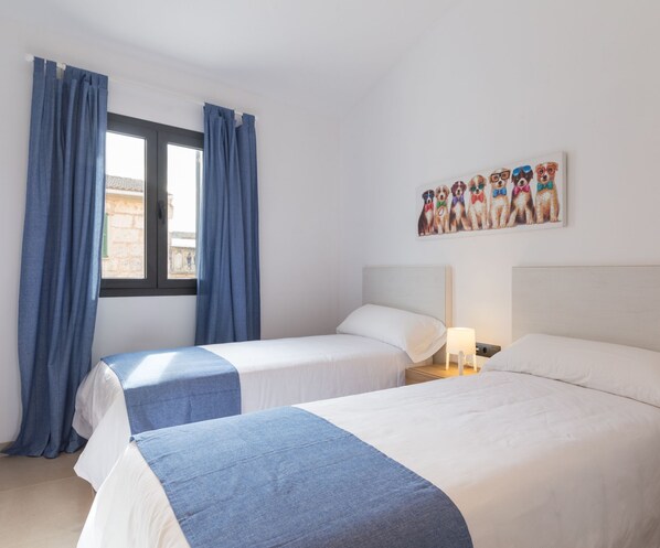 Une chambre d’hôtel avec deux lits, une fenêtre avec des rideaux, un mur orné d’un tableau sur le thème des chiens et une table de chevet avec une lampe.