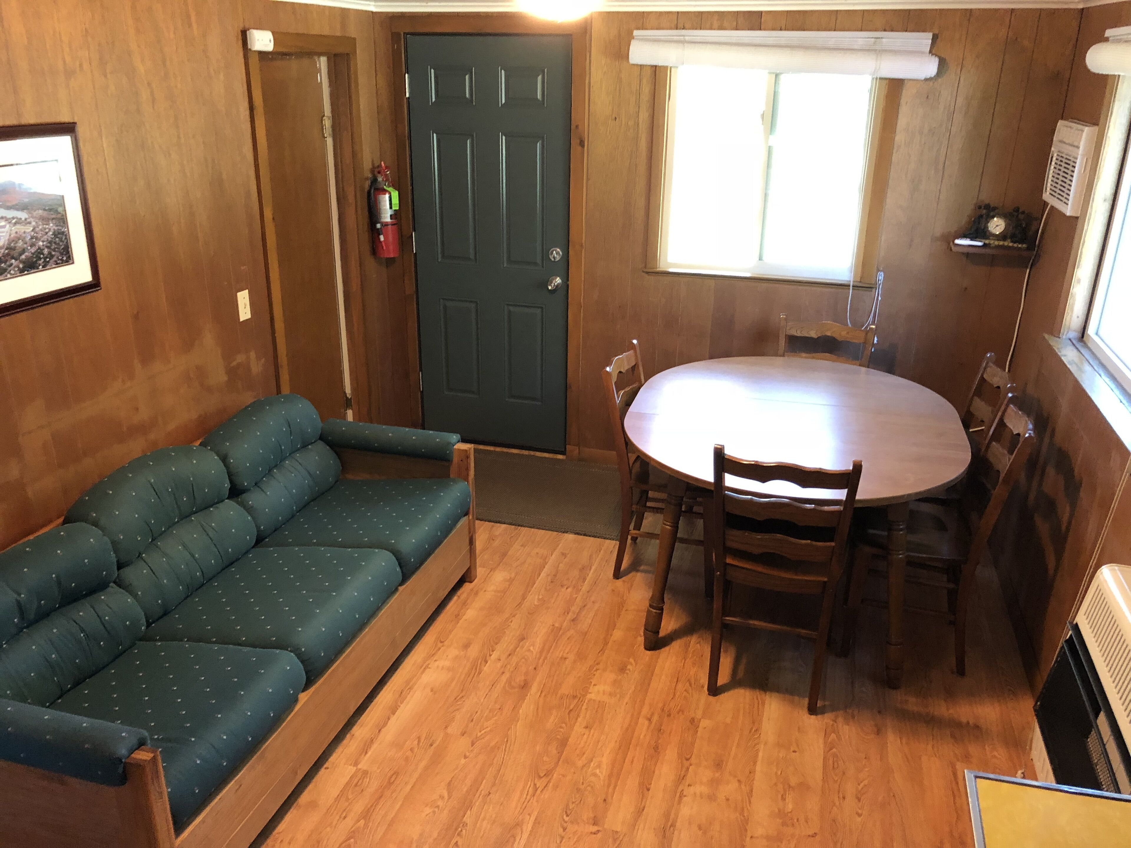 A compact room with a wooden floor, a green sofa, a round dining table with chairs, a dark door, a window with blinds, and a small shelf with a clock.