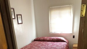 Quarto com piso de madeira, cama com colcha florida, mesa de cabeceira e janela com persianas.