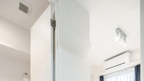 Ein modernes Hotelzimmer mit einer weißen Toilette, einem Bett mit weißer Bettwäsche, einem Fenster mit blauen Vorhängen und einer Klimaanlage an der Wand.