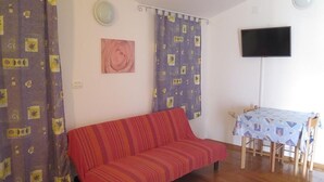 Una sala de estar con un sofá rojo, una mesa con mantel estampado y un televisor montado en la pared.