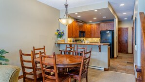 A dining area with a wooden table and chairs, a kitchen with wooden cabinets, and a refrigerator. - Excellent multi level 2 bedroom + den townhouse, close to Morrisey lift (Sun Peaks)