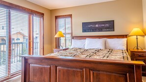 A bedroom with a wooden bed, two bedside lamps, a nightstand, and a cityscape painting on the wall. - Excellent multi level 2 bedroom + den townhouse, close to Morrisey lift (Sun Peaks)