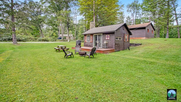 A cabin with a porch, picnic table, and chairs, surrounded by a grassy area and trees.