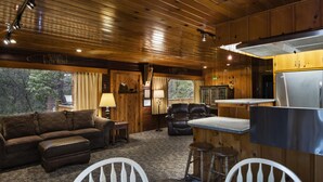 Ein gemütliches Blockhauss-Interieur mit Holzbalkendecke, einem Wohnbereich mit Sofa und Sesseln, einer Küche mit Kühlschrank und einer Bar.