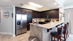 A modern kitchen with dark wood cabinets, stainless steel appliances, a granite countertop, and a bar area with bar stools. - Phoenix Orange Beach 2006 by Liquid Life (Orange Beach)