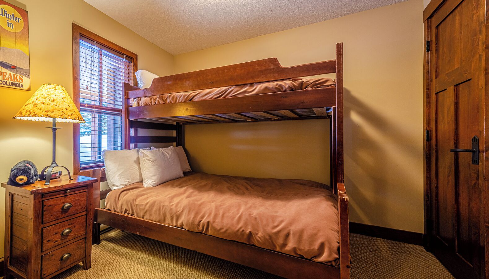 Een stapelbed met houten frame, een nachtkastje met lades, een lamp en een raam met jaloezieën.
