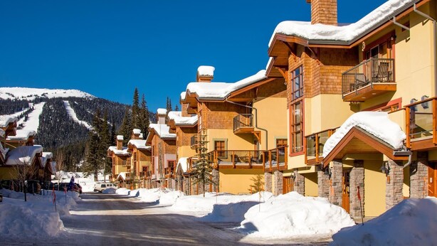 A row of log cabin-style buildings with snow-covered roofs and a mountain backdrop. - Exceptional multi level 2 bedroom +den townhouse with easy access to nearby runs and lift (Sun Peaks)