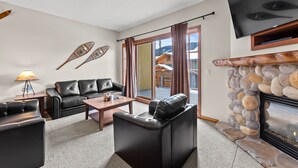 A modern living room with a stone fireplace, leather sofas, a flat-screen TV, and large windows offering a view of a cabin. - Exceptional multi level 2 bedroom +den townhouse with easy access to nearby runs and lift (Sun Peaks)
