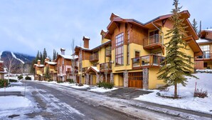 A row of multi-story buildings with balconies, a snow-covered street, and a mountainous backdrop. - Exceptional multi level 2 bedroom +den townhouse with easy access to nearby runs and lift (Sun Peaks)