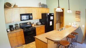 A compact kitchen with wooden cabinets, a black stove, a microwave, and a refrigerator. There are three stools at the bar counter. - Upper floor 2 bedroom condo with courtyard view and easy access to ski runs (Sun Peaks)
