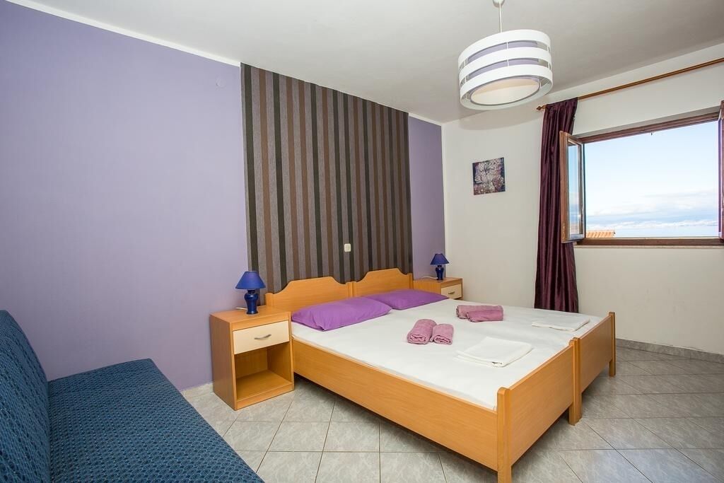 Ein Schlafzimmer mit einem hölzernen Bett, violetter Bettwäsche, einer gestreiften Wand, einem Fenster mit Vorhängen und einem blauen Sessel.