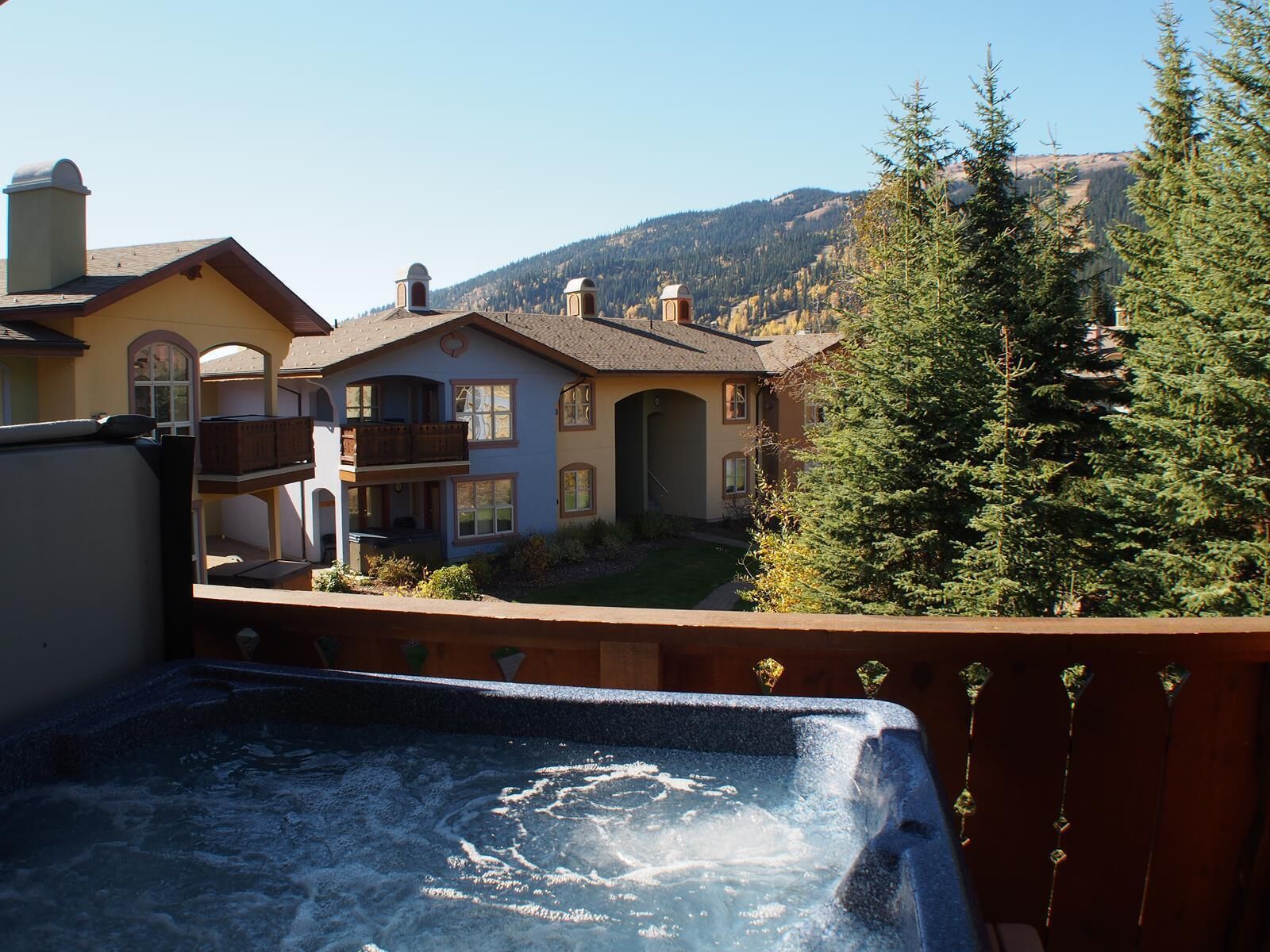 Ein Whirlpool mit einer hölzernen Terrasse vor einem Haus mit Balkon, umgeben von Bäumen und einer Bergkulisse.
