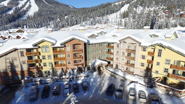 A snow-covered resort with multi-story buildings, a parking lot, and a mountainous backdrop.