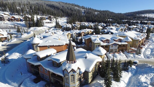 A snow-covered resort with multiple buildings, including a prominent tower, surrounded by trees and a clear sky.