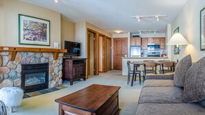 A living room with a stone fireplace, a television, and a sofa. There is a kitchen in the background with wooden cabinets and a dining area. - 1 bedroom apartment in village centre, ski in/out (Sun Peaks)