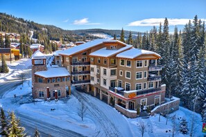 Un hotel de varios pisos con techo nevado, múltiples balcones y una pista de esquí al fondo.