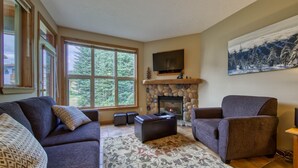 A living room with a stone fireplace, a large window, and a painting of a snowy landscape. - Ground floor 2 bedroom condo with easy access to ski runs (Sun Peaks)