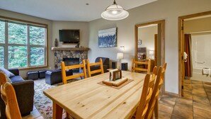 A dining area with a wooden table and chairs, a fireplace, a television mounted above it, and a large window offering a view of the outdoors. - Ground floor 2 bedroom condo with easy access to ski runs (Sun Peaks)