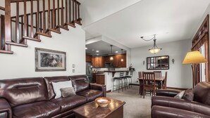 A spacious living room with a brown leather sofa, a wooden coffee table, and a dining area with wooden chairs and a kitchen in the background. - Spacious multi level 3 bedroom + den townhouse, close to Morrisey lift (Sun Peaks)