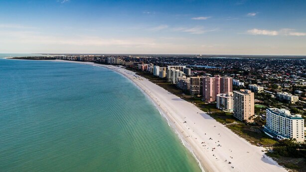 Vue aérienne d’une ville côtière avec une longue plage de sable, de hauts immeubles et un ciel bleu dégagé.