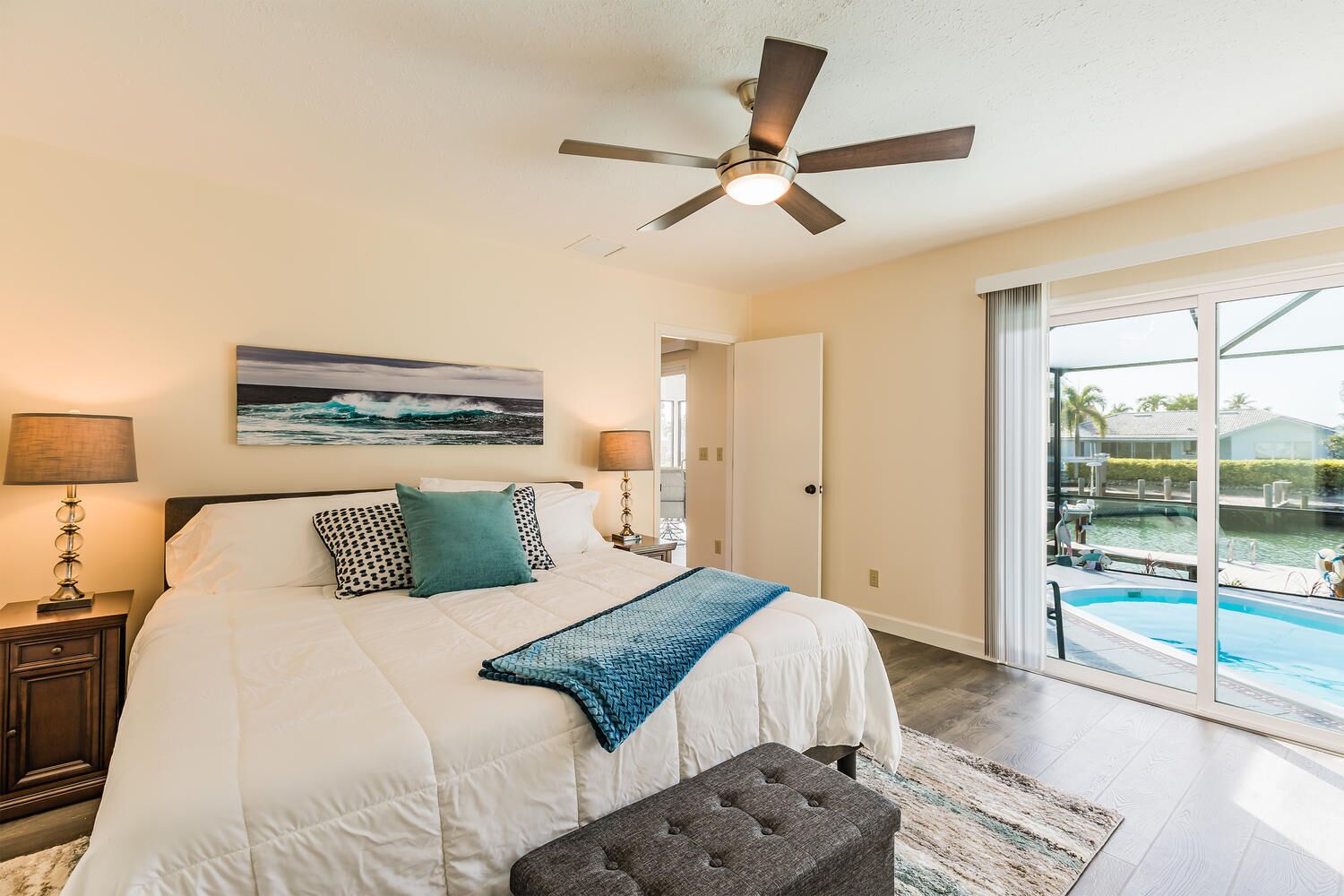 A bedroom with a bed, a ceiling fan, a nightstand, a lamp, a painting, and a view of a swimming pool.
