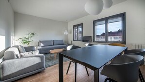 Una sala de estar moderna con un sofá gris, una mesa de centro y una mesa de comedor con sillas. Hay un ventanal con vistas a edificios.
