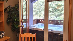 A wooden chair in front of a glass door leading to a hot tub area with a view of a forested landscape. - Cozy Condo Close to Village | Access to Ski-Out Trail | Private Hot Tub | Fireplace (Sun Peaks)