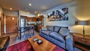 A modern living room with a grey sofa, a coffee table, and a dining area with a bar stool. - Modern 1 bedroom apartment in village centre (Sun Peaks)