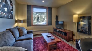 A modern living room with a sofa, a coffee table, a TV on a wooden stand, a large window with a view of trees, and a red carpet. - Modern 1 bedroom apartment in village centre (Sun Peaks)