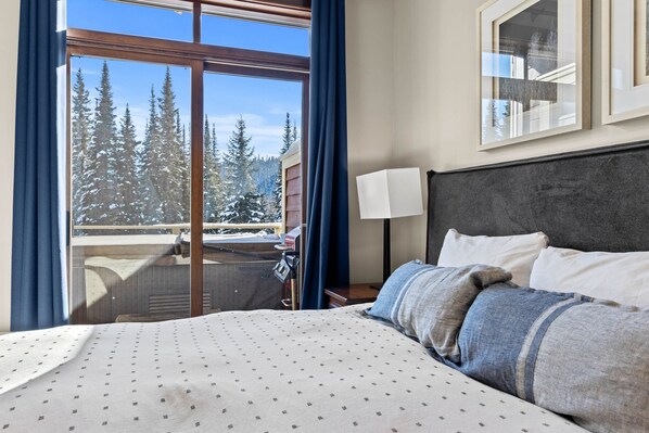 Relax in a spacious room with a large bed, wooden headboard, and a bedside table featuring a lamp. Enjoy a view of snow-covered trees from the balcony.