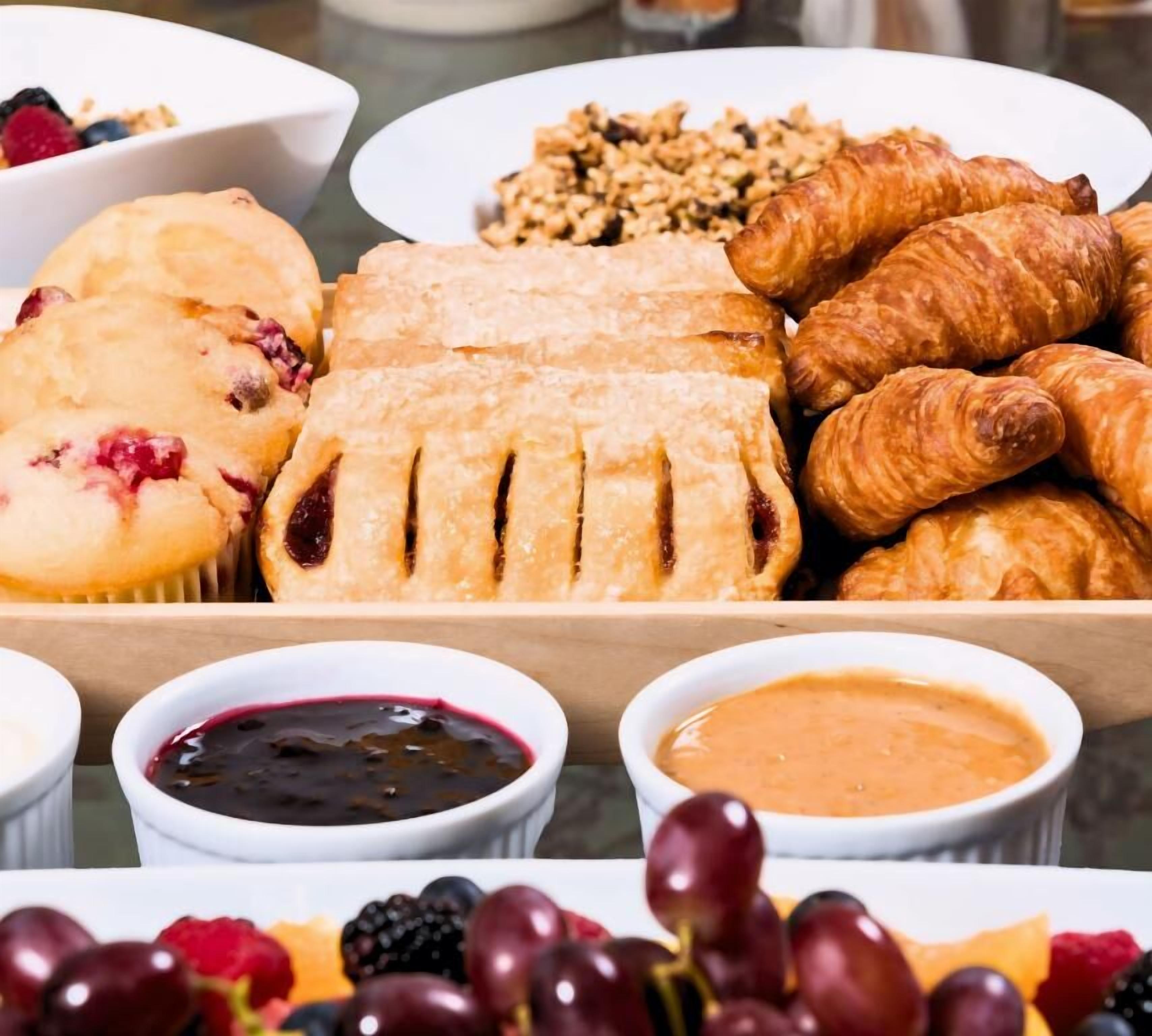 Diversas opciones de pastelería, incluyendo tartas, croissants y pan, junto con recipientes de mermelada y crema de cacahuete.