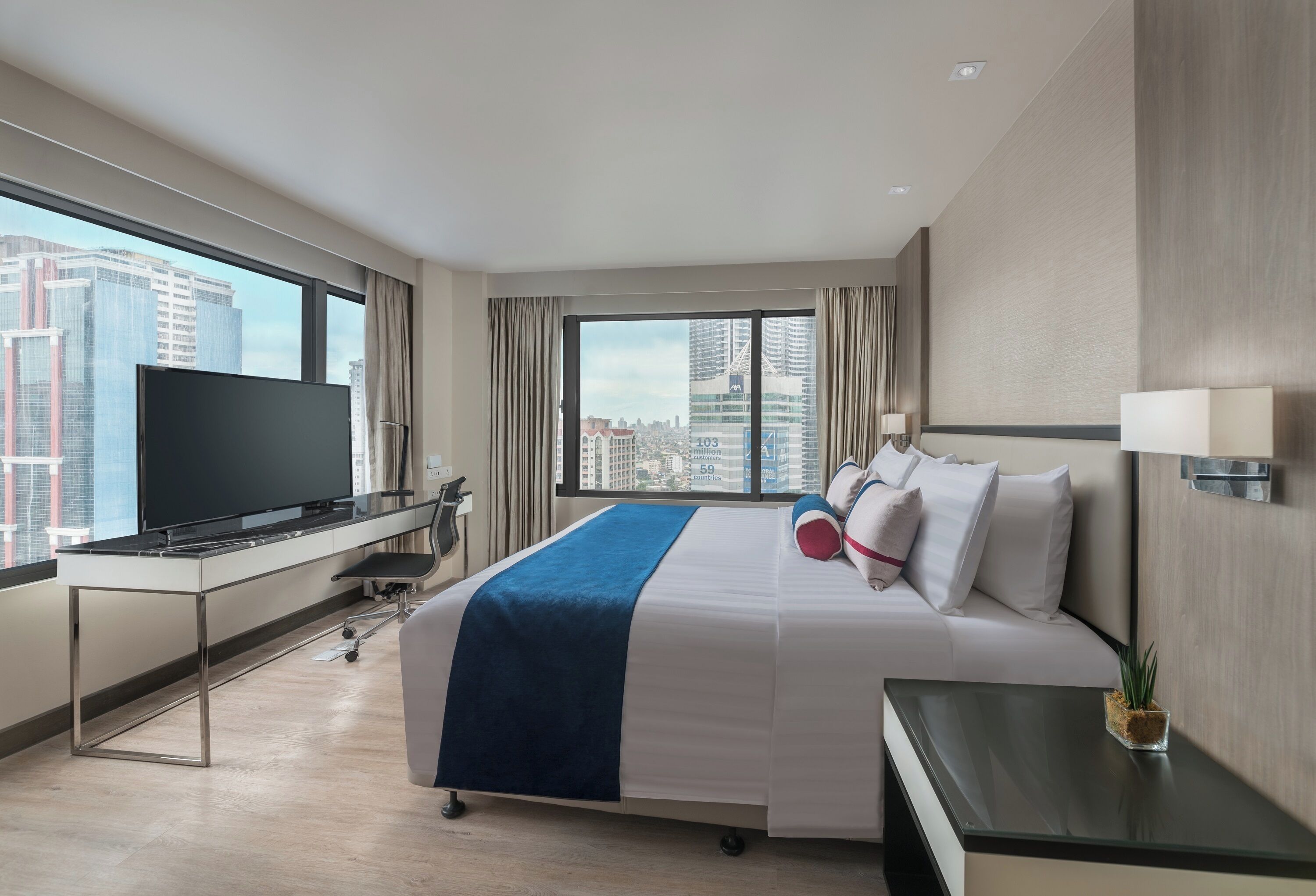 A hotel room with a large bed, a desk with a flat-screen TV, a small table, and a view of the cityscape through large windows.