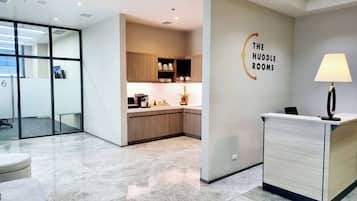 A modern reception area with a kitchen in the background, a reception desk, and a wall sign reading "The Huddle Rooms".