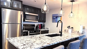 A modern kitchen with a stainless steel refrigerator, a microwave, an oven, and a sink with a black faucet. There are two pendant lights with a geometric design. The kitchen island has a marble-like countertop. - Best True Ski in Out Rare slopeside @ 5 lifts Pool/Spa/Gym/WD/Parking Near Main! (Park City)