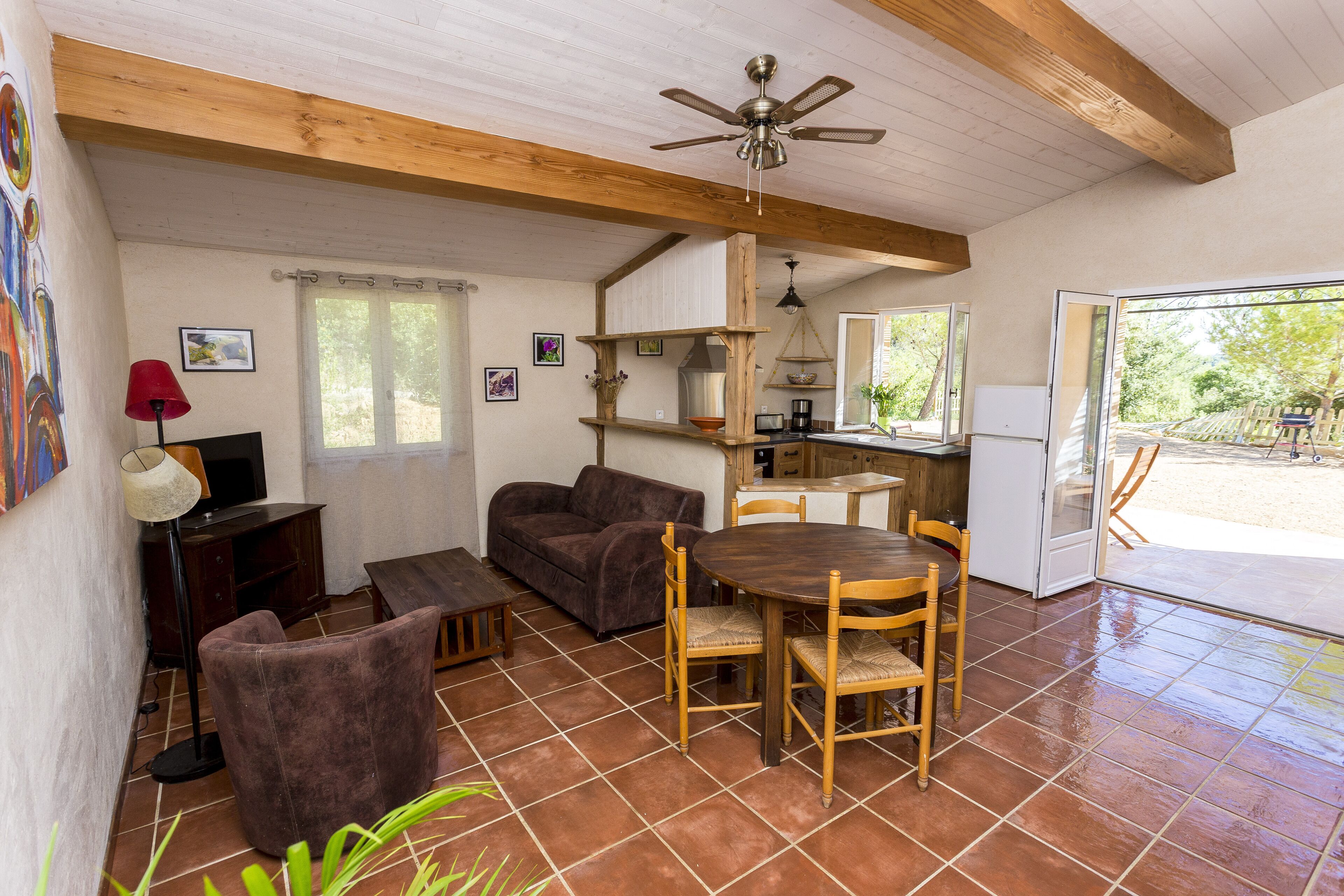 A cozy living area with a sofa, a wooden table, and chairs. There is a kitchen area with a refrigerator and a sink. The room has a tiled floor and a ceiling fan.