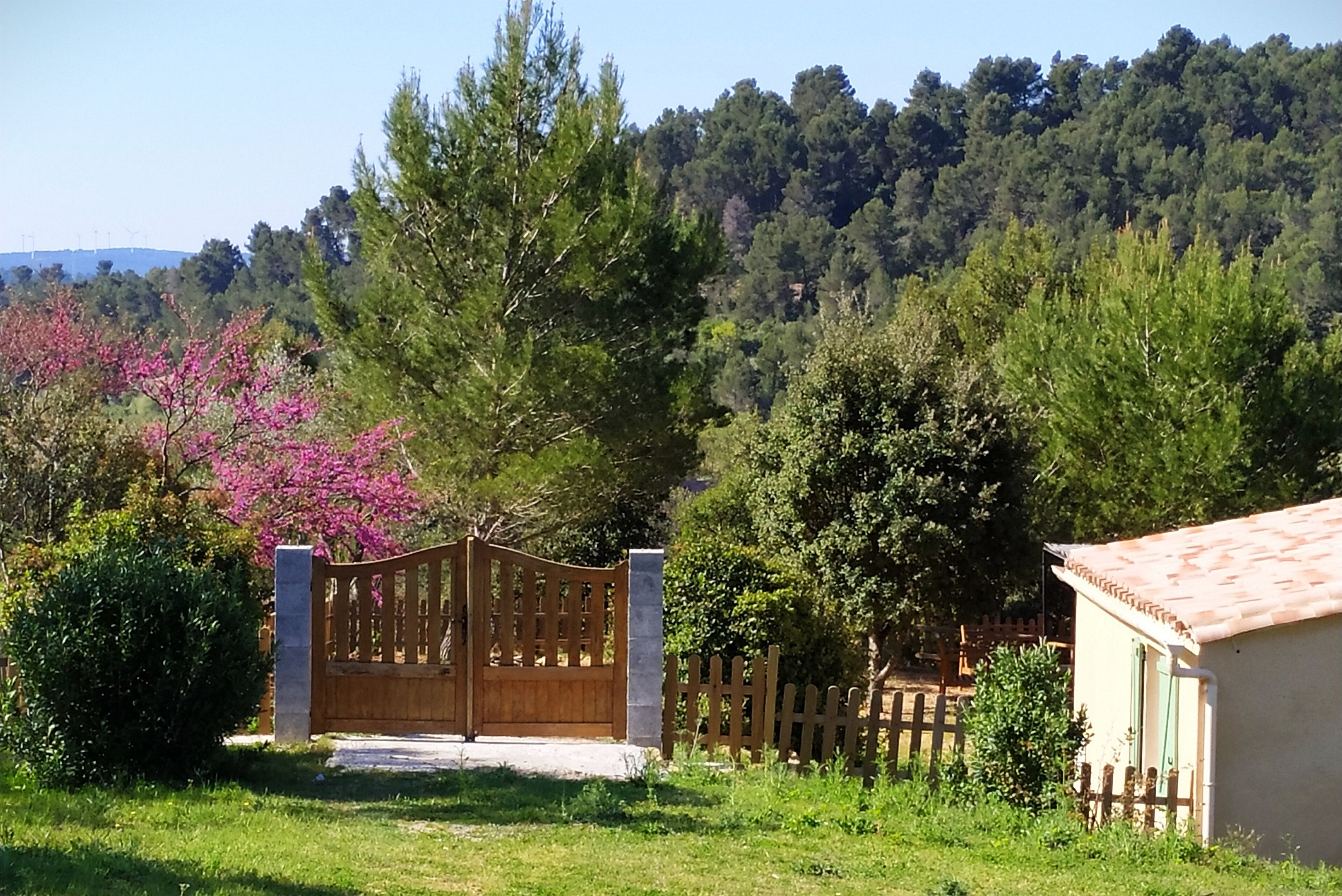 Une entrée fermée par un portail en bois, entourée d’une végétation luxuriante et avec une colline en arrière-plan.