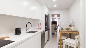 Cocina moderna con gabinetes blancos, un grifo negro y una cafetera. Hay un comedor con una mesa y sillas, y un televisor al fondo.