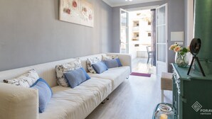Una sala de estar moderna con un sofá blanco, cojines azules, un cuadro abstracto grande, una lámpara de araña y vistas al balcón exterior.