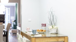 Un comedor con una mesa de vidrio, sillas blancas, un plato de croissants, una cesta de flores y una taza.