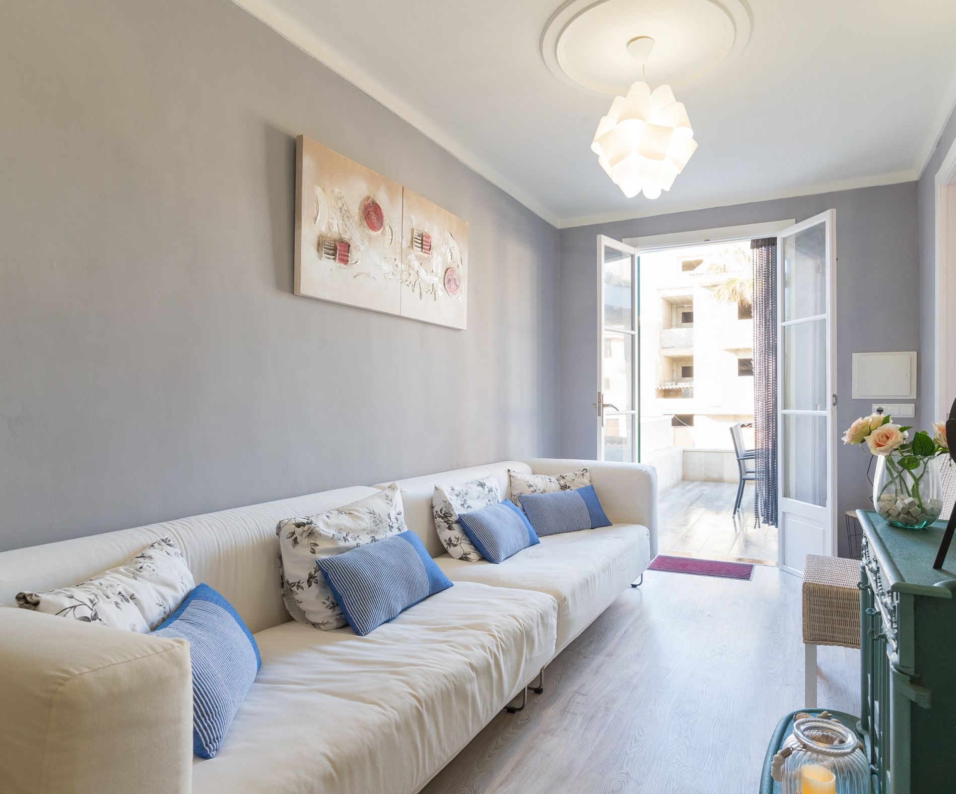 Una sala de estar moderna con un sofá blanco, cojines azules, un cuadro abstracto grande, una lámpara de araña y vistas al balcón exterior.