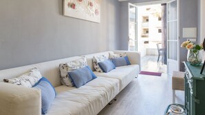 A modern living room with a white sofa, blue cushions, a large abstract painting, a chandelier, and a view of the outside balcony.